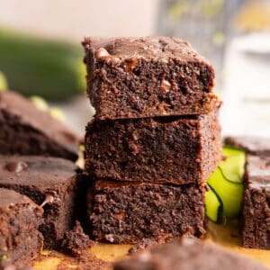 Three zucchini brownies in a stack surrounded by ingredients and more brownies.