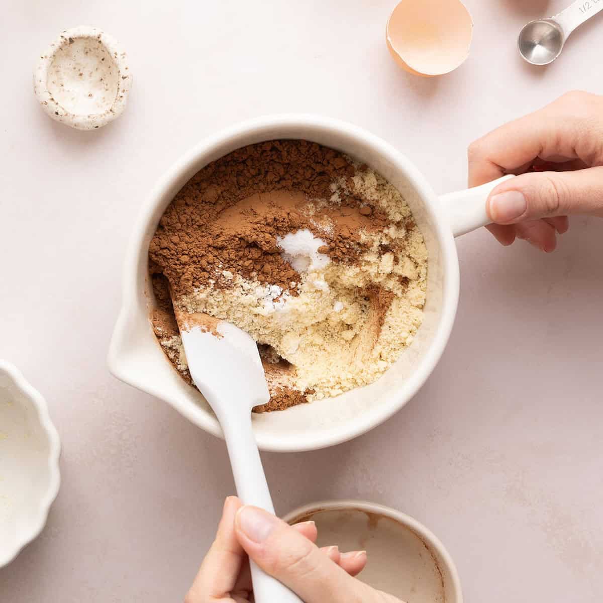 Woman mixing almond flour, cocoa powder, salt, and baking soda in a bowl.