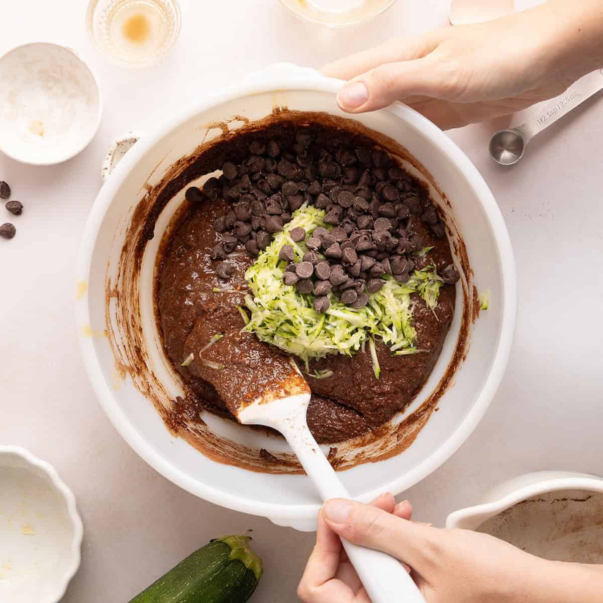 Woman using a rubber spatula to fold grated zucchini into a bowl of brownie batter.