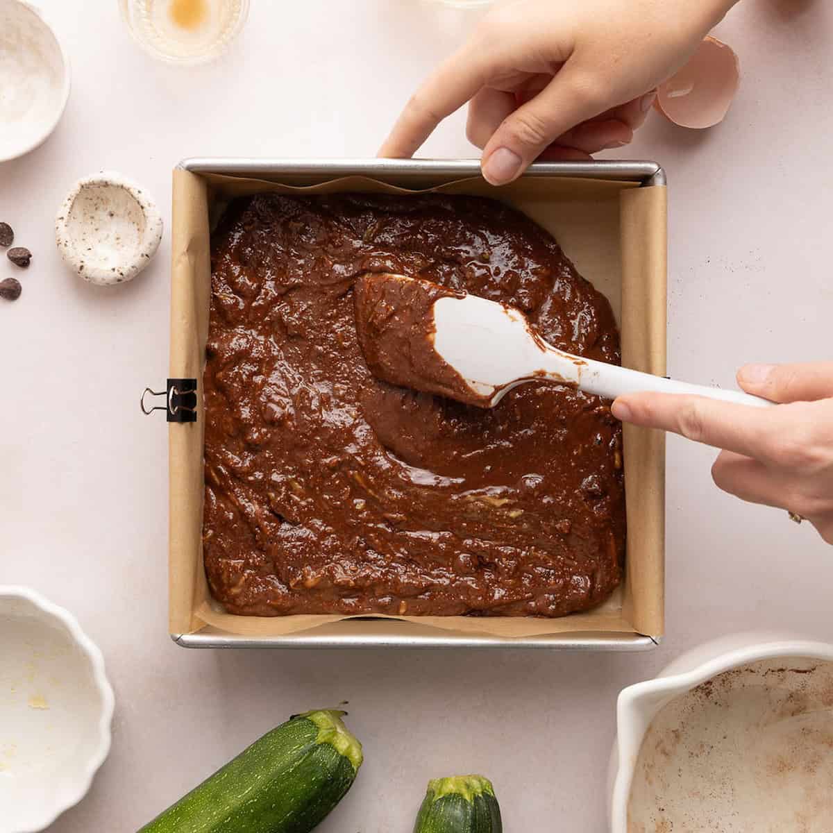 Woman spreading zucchini brownie batter into a square baking pan with parchment paper.