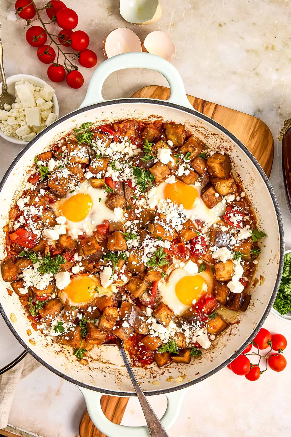 Eggplant shakshuka with four poached eggs in a large skillet on a board surrounded by ingredients.