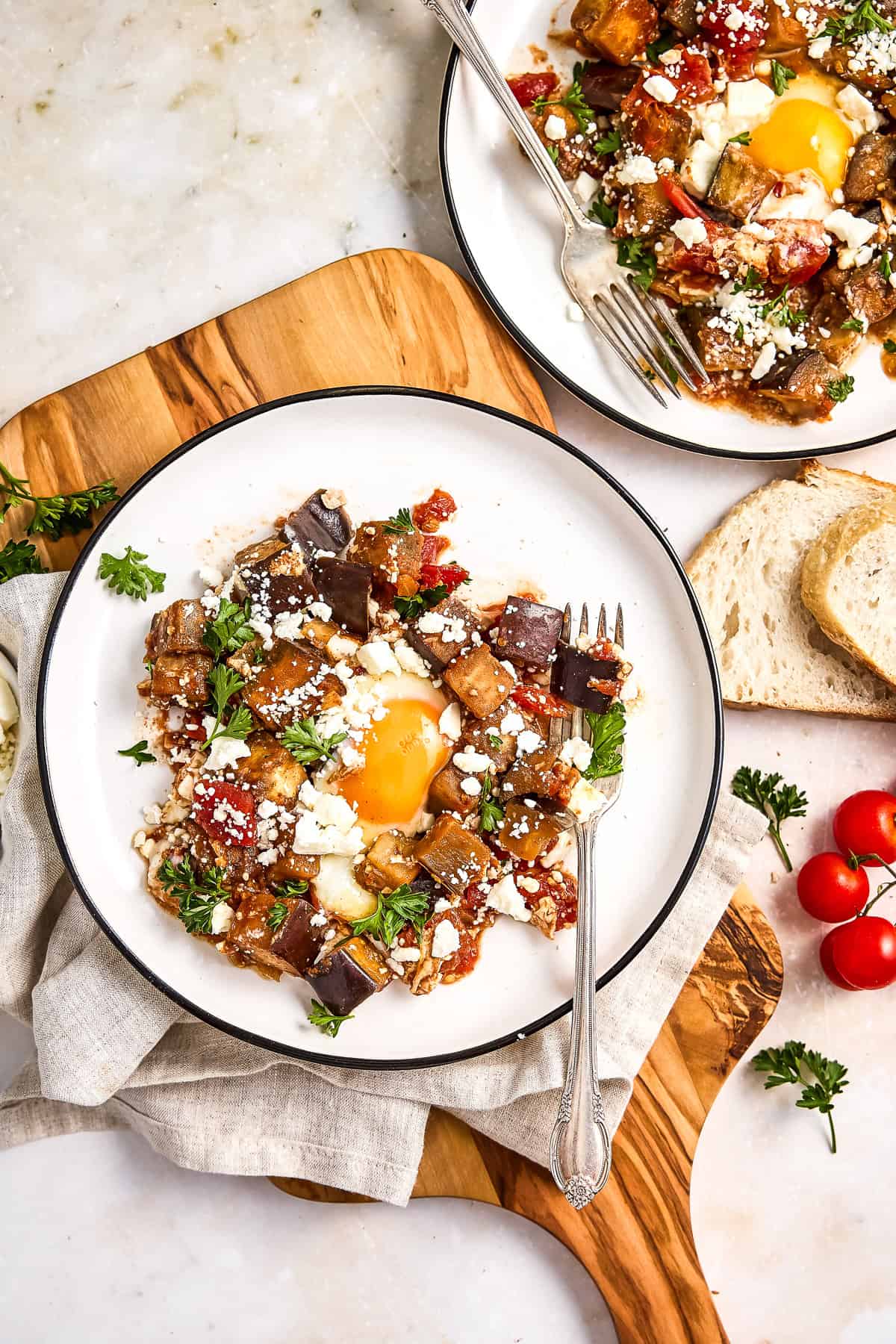 Eggplant and tomato shakshuka on serving plates surrounded by bread and ingredients.