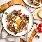 Eggplant and tomato shakshuka on serving plates surrounded by bread and ingredients.