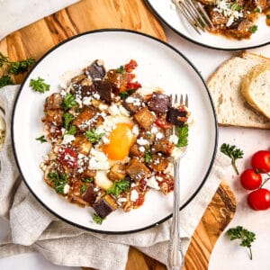 Eggplant and tomato shakshuka on serving plates surrounded by bread and ingredients.