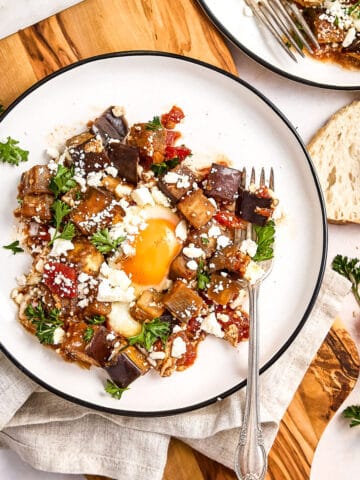 Eggplant and tomato shakshuka on serving plates surrounded by bread and ingredients.
