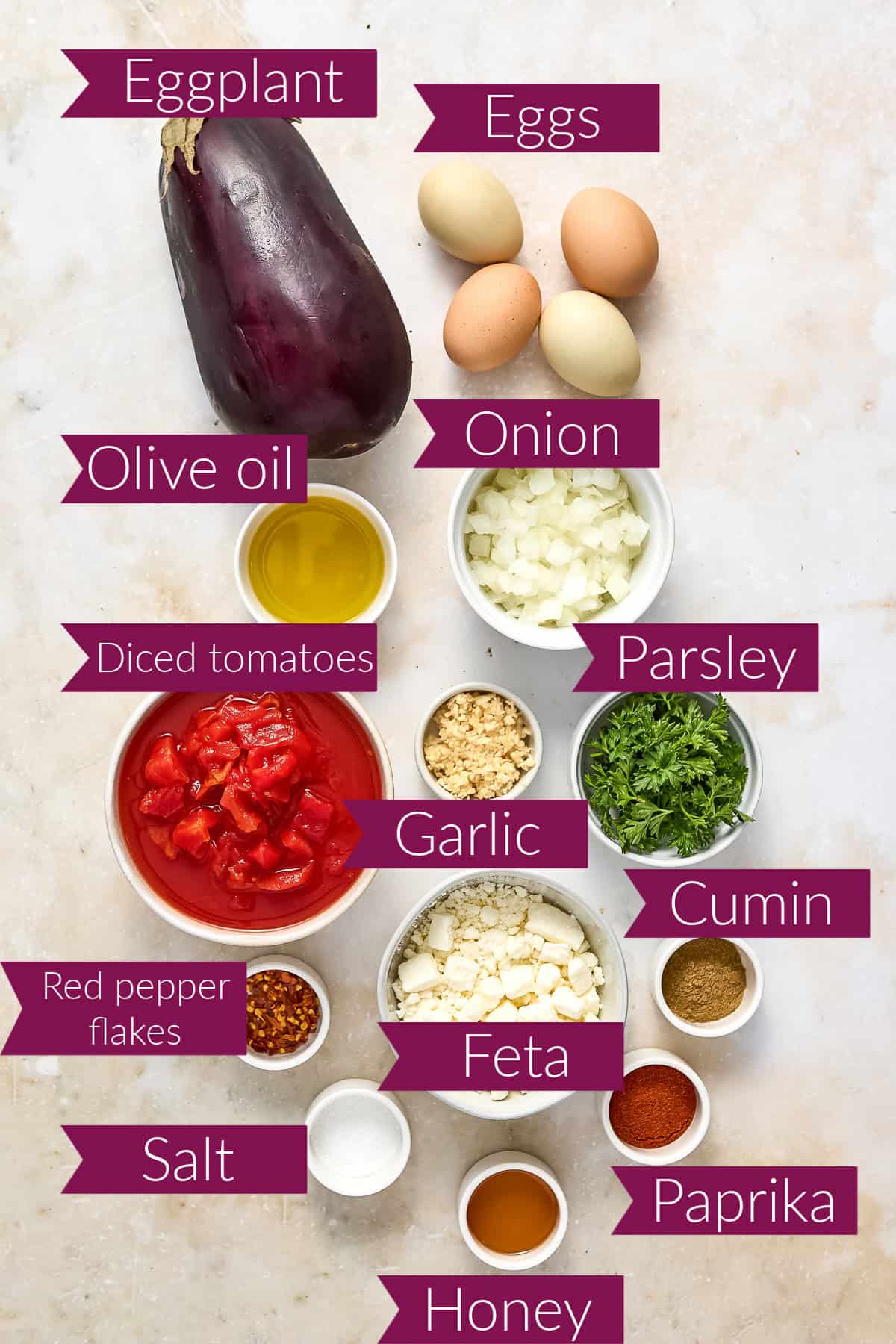 Eggplant, eggs, oil, onion, tomatoes, garlic, parsley, spices, and feta cheese on a counter with labels.