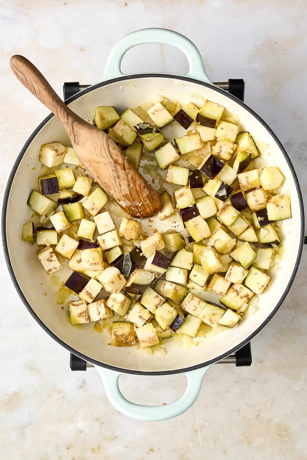 Eggplant and onion cooking in a large skillet with a wooden spoon.