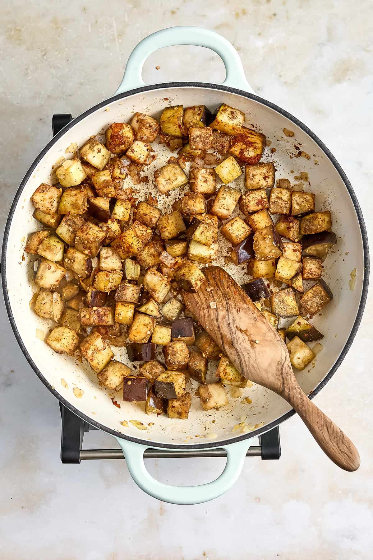 Eggplant cooked down with spices in a large skillet with a wooden spoon.