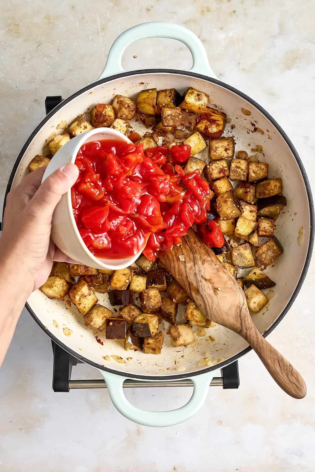 Woman adding diced tomatoes to a skillet with eggplant, onion, and spices.