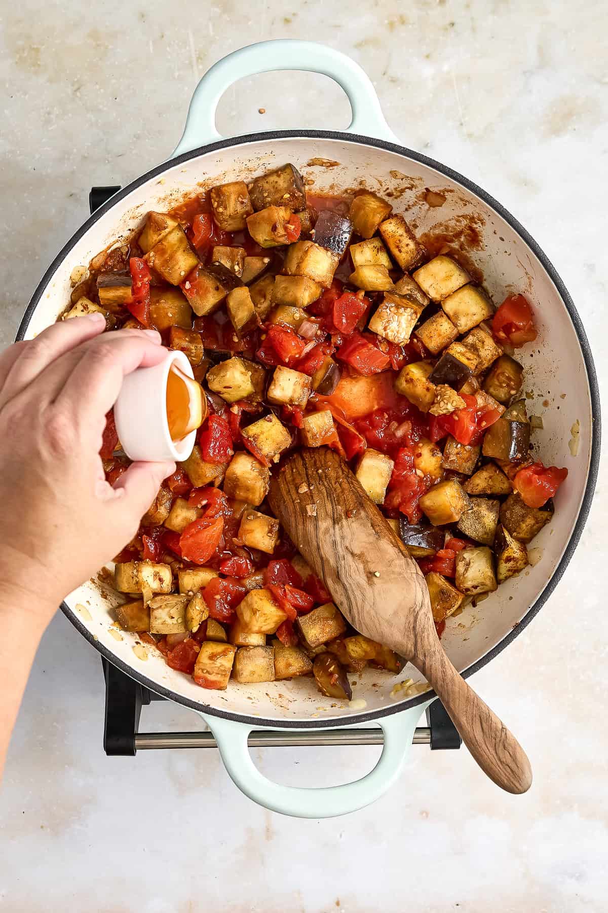 Woman adding honey to a skillet of eggplant shakshuka ingredients cooking.