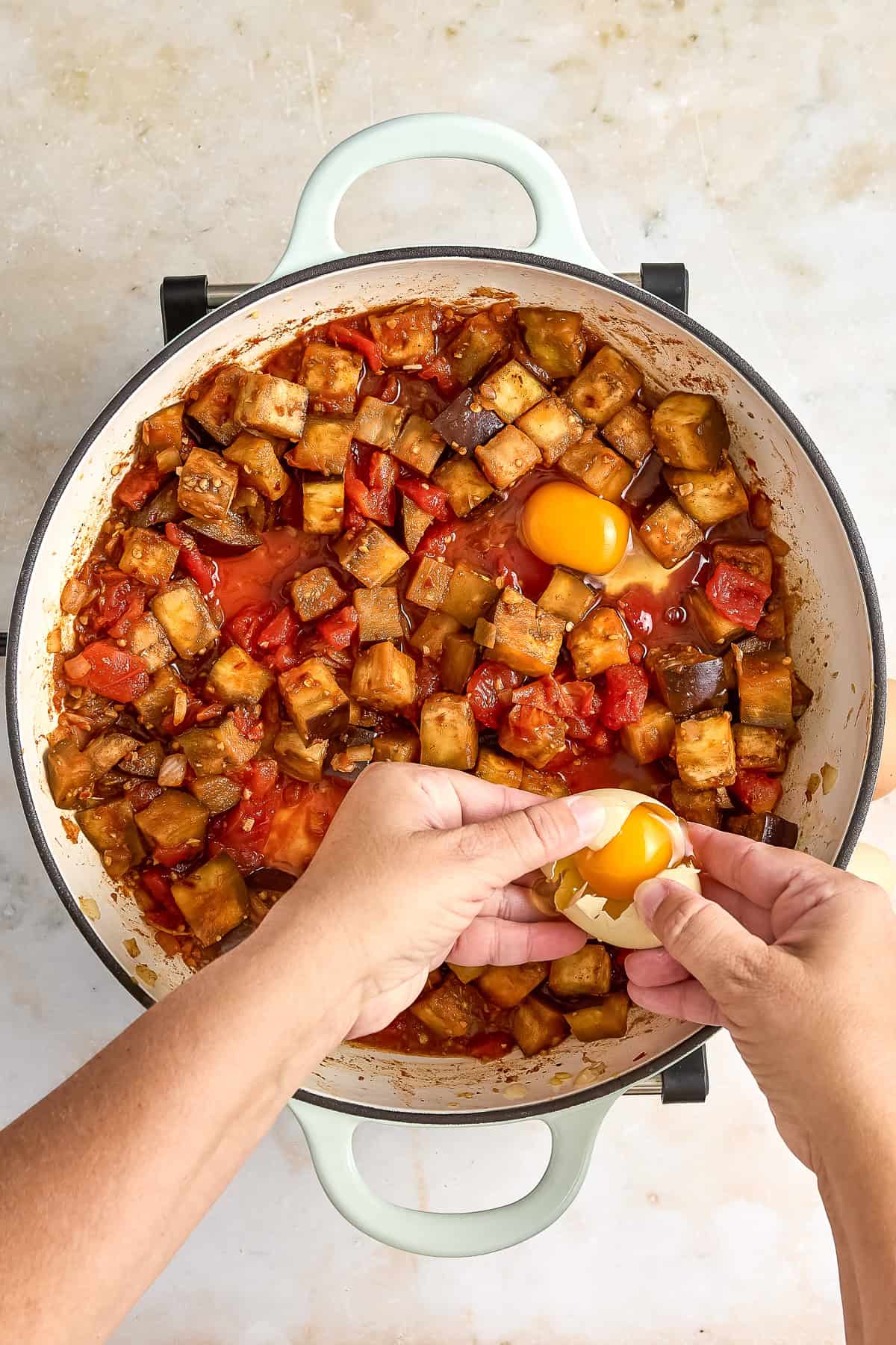 Woman cracking eggs into a skillet with eggplant shakshuka cooking.