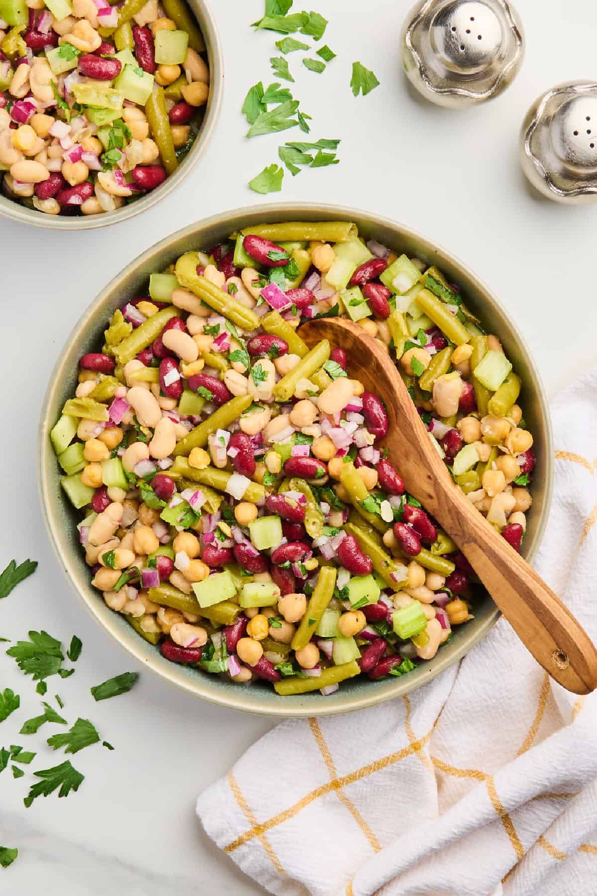 Bowl of four bean salad with a wooden spoon next to napkin, herbs, salt, and pepper.