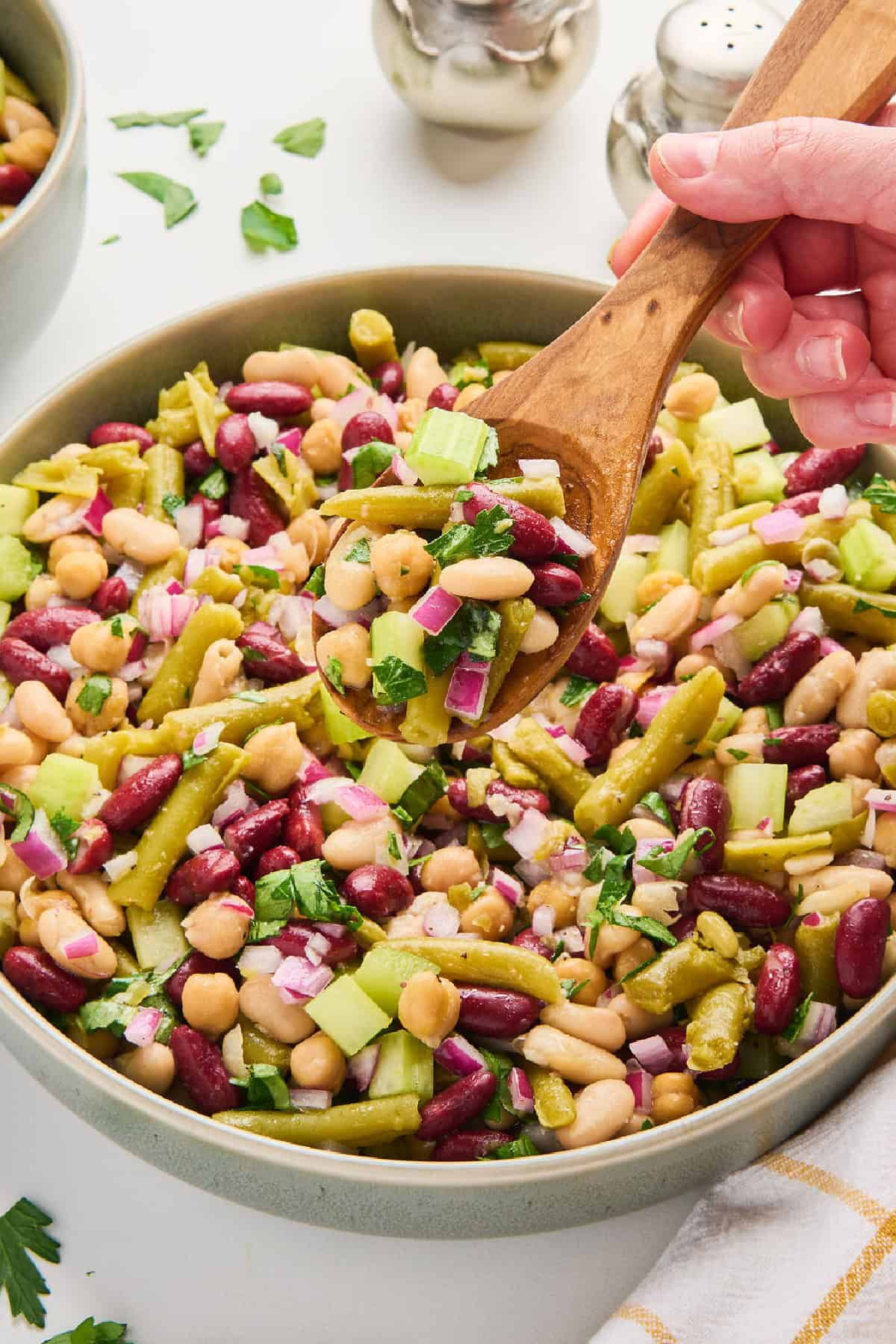 Woman holding a scoop of four bean salad in a wooden spoon over a bowl.