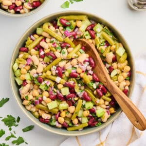 Bowl of four bean salad with a wooden spoon next to napkin, herbs, salt, and pepper.