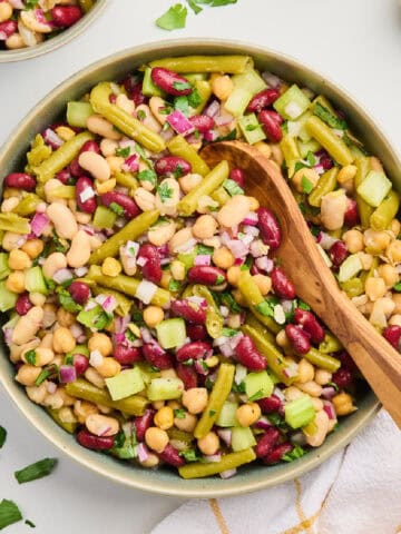Bowl of four bean salad with a wooden spoon next to napkin, herbs, salt, and pepper.