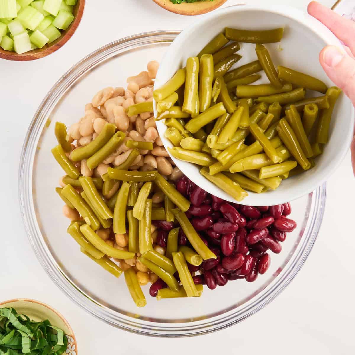 Woman adding green beans to glass bowl with kidney beans and white beans.