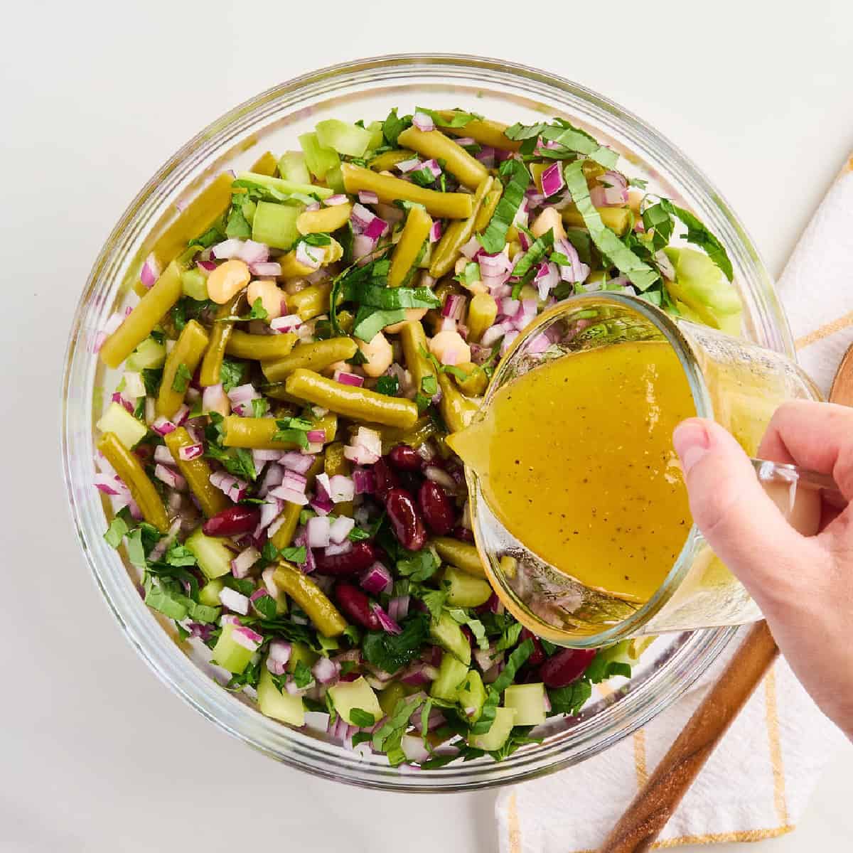 Woman pouring apple cider vinaigrette over bean salad in a glass bowl.