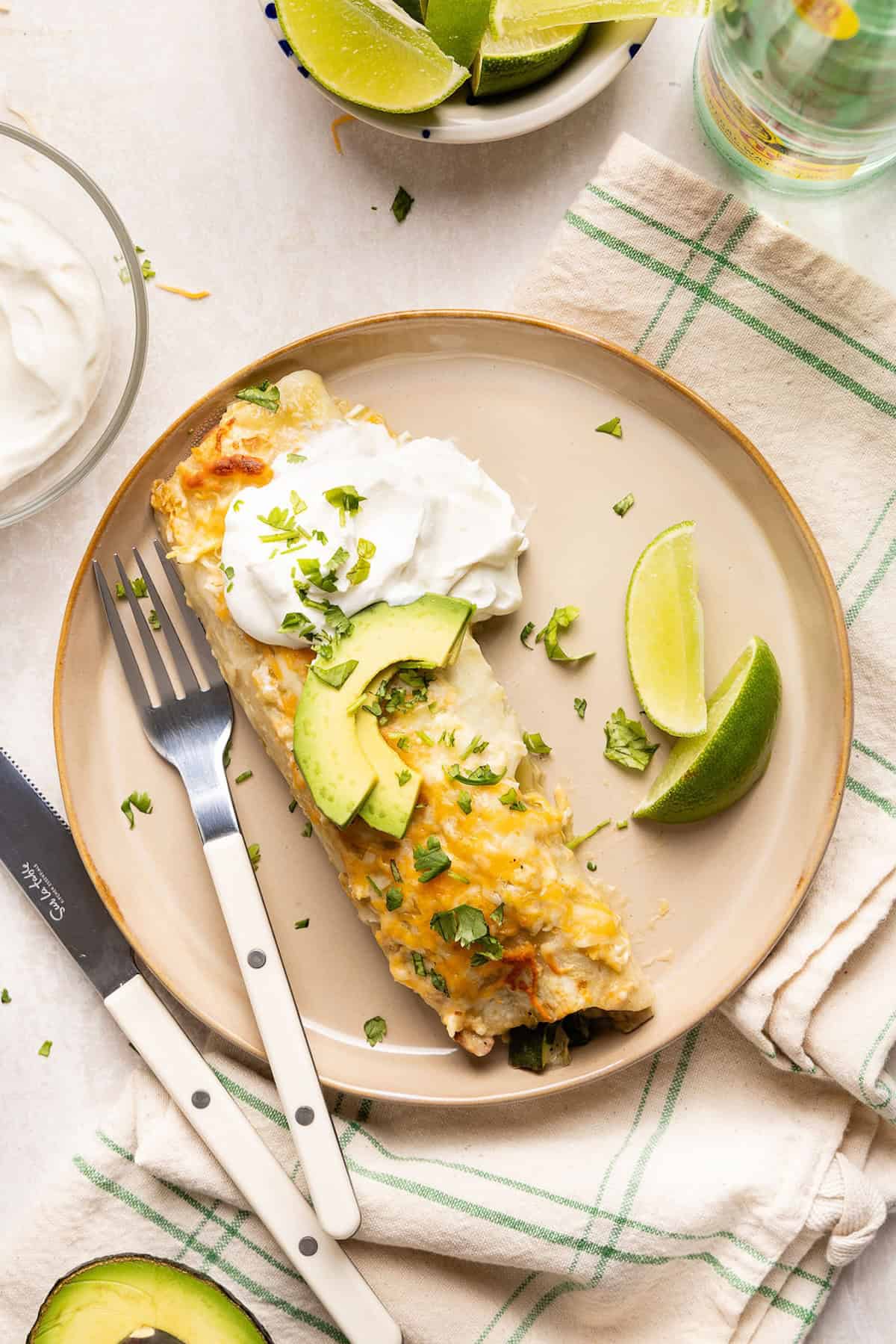 One enchilada on a plate with sour cream, avocado, lime, and a fork.