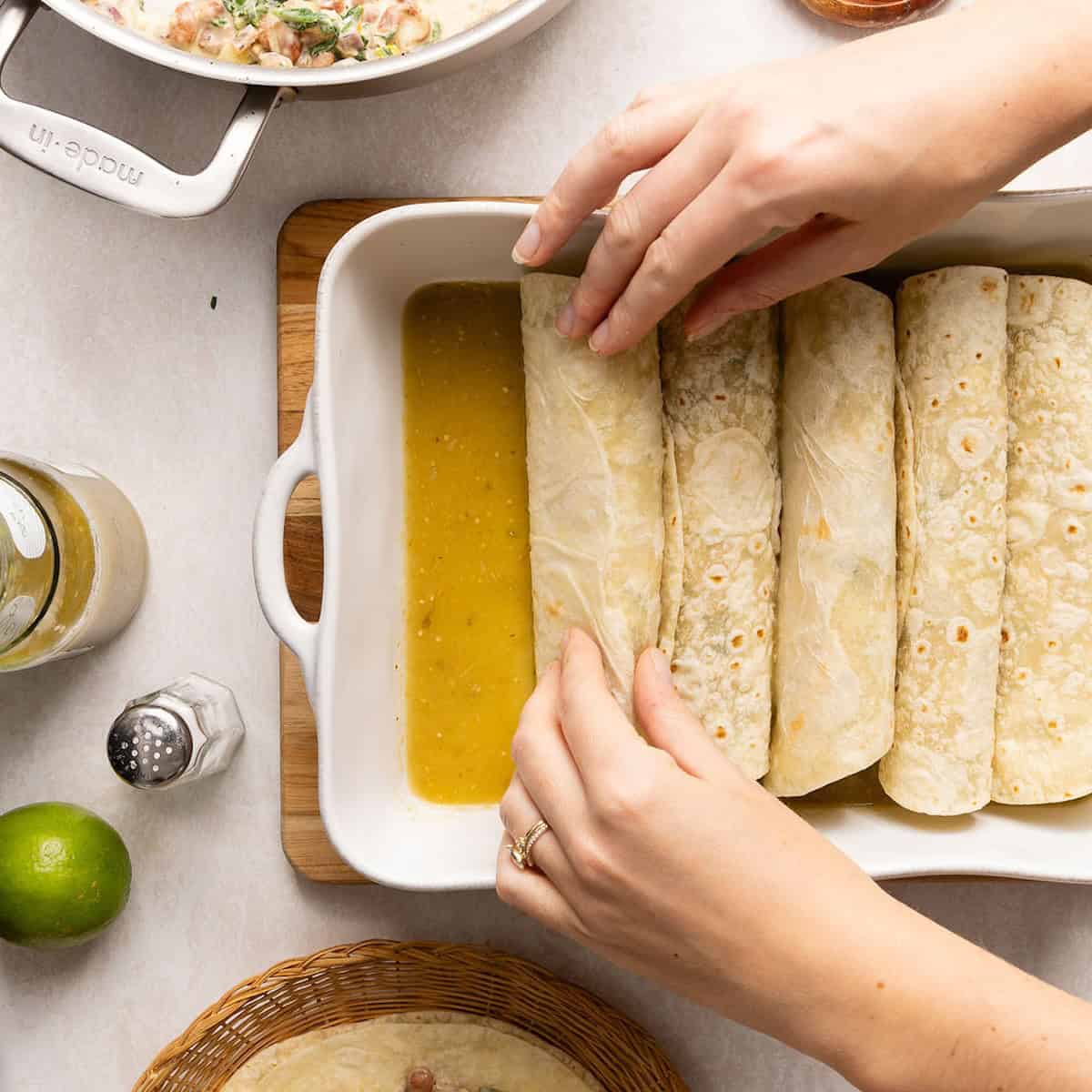 Woman placing rolled enchiladas into a baking dish with salsa verde on the bottom.
