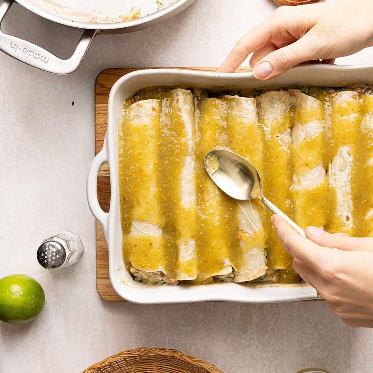 Woman spreading salsa verde on top of enchiladas in a baking dish with a spoon.