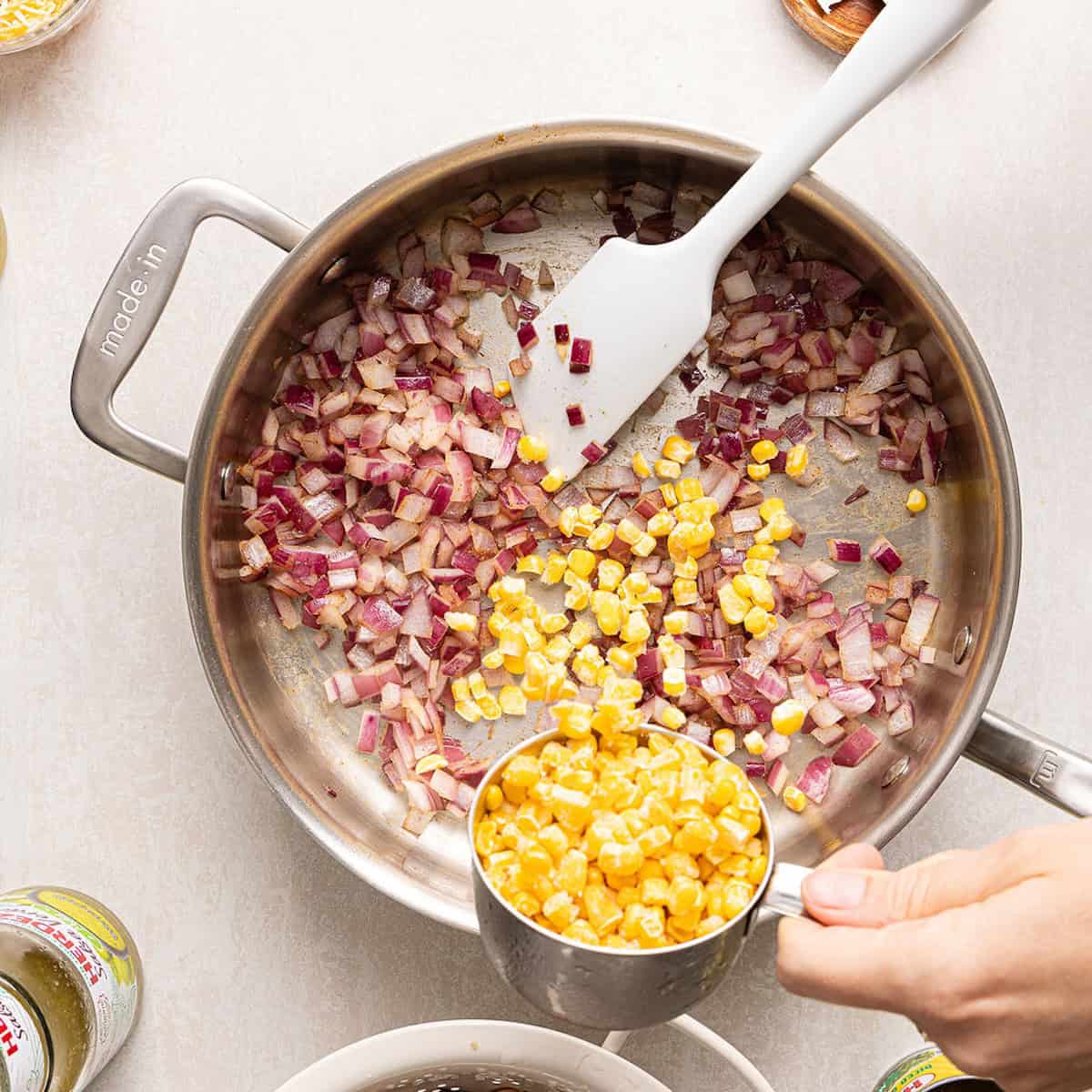 Woman adding frozen corn to a skillet with cooked red onion.