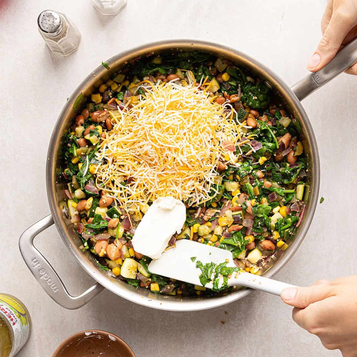 Shredded Mexican cheese and cream cheese added to a skillet with cooked vegetables.