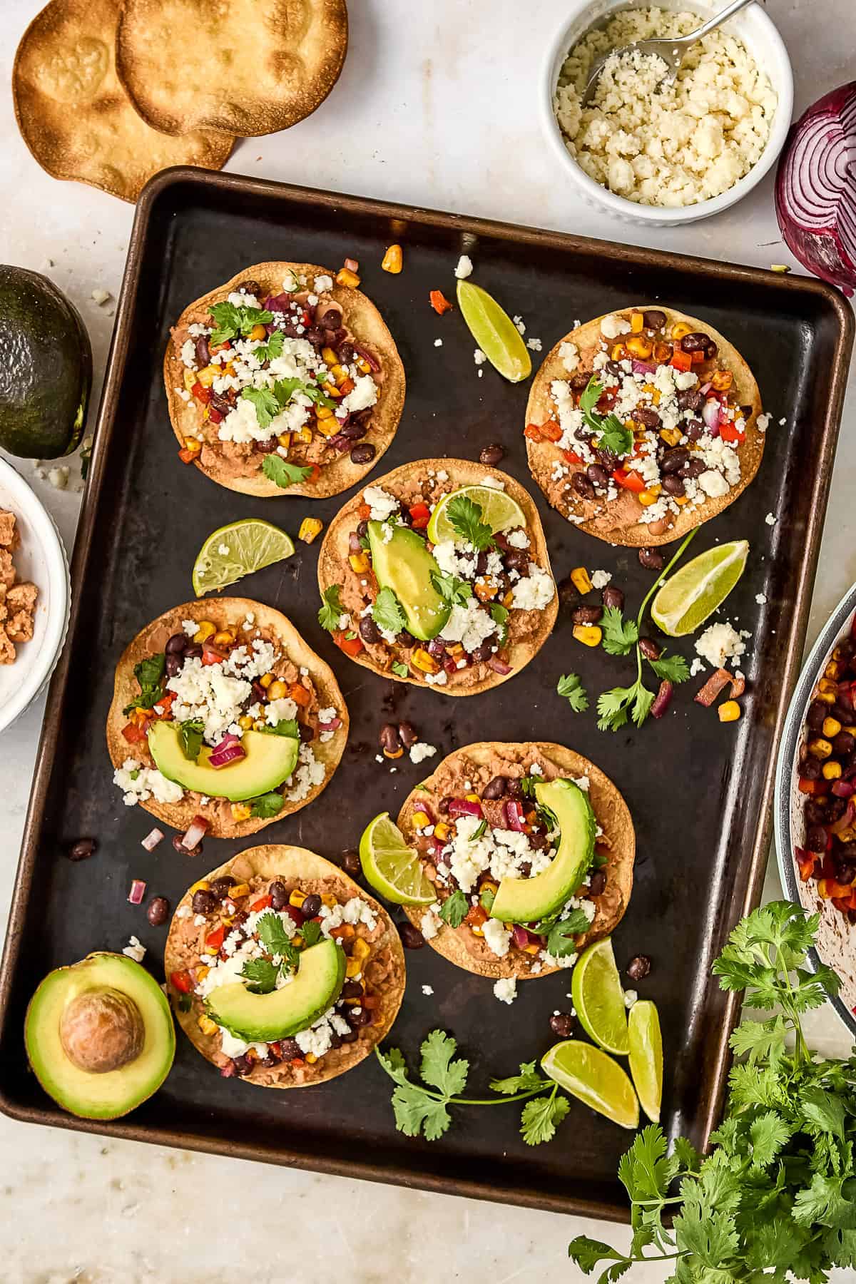 Five vegetarian tostadas on a baking sheet with avocado and lime wedges.