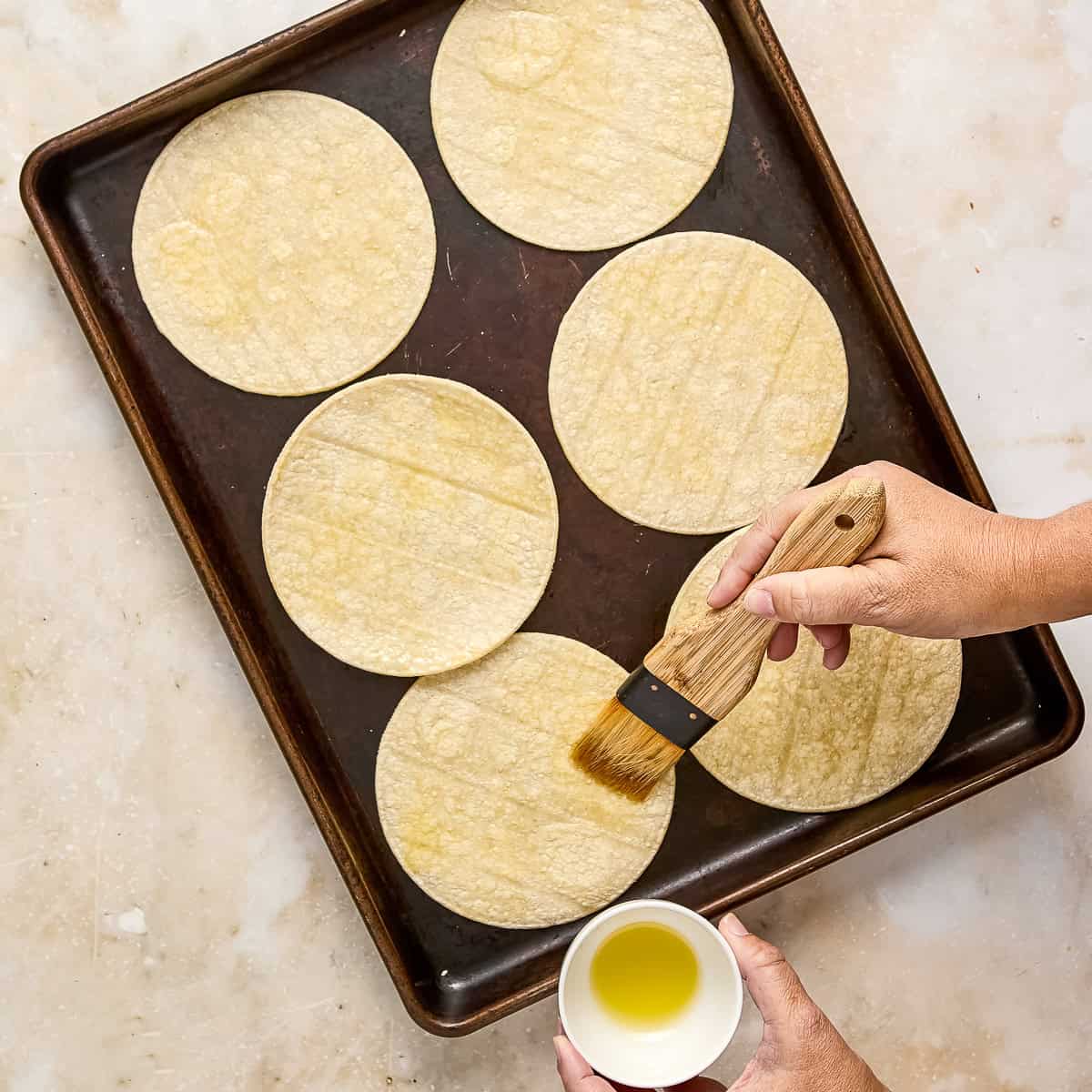 Woman brushing oil on corn tortillas on a baking sheet.