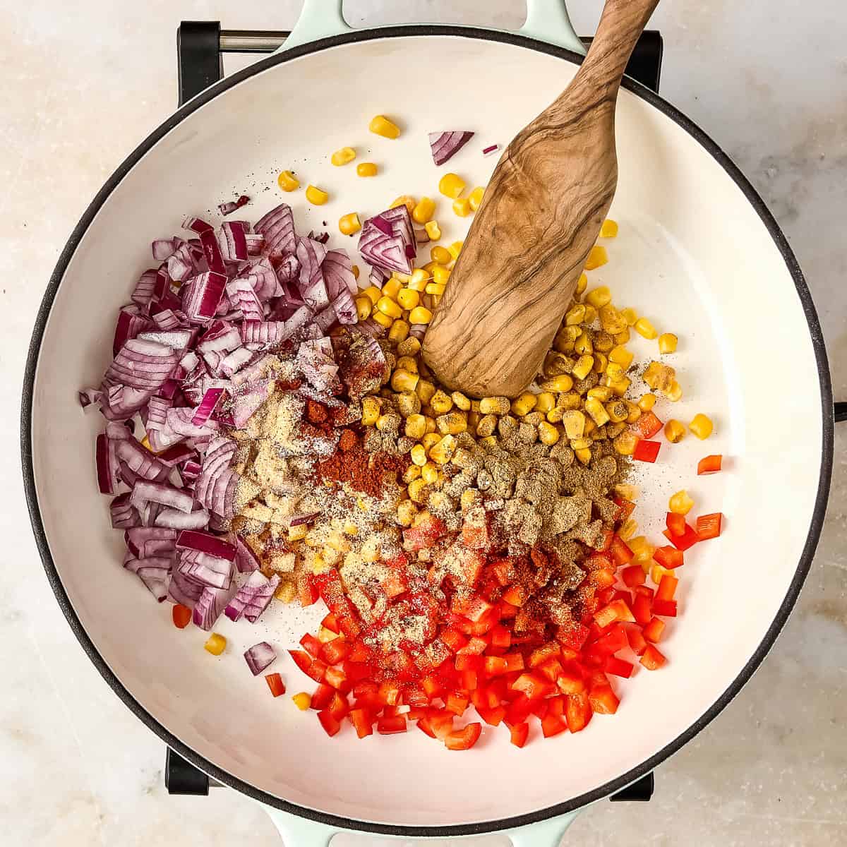 Red onion, peppers, corn, and seasonings in a skillet with a wooden spoon before cooking.
