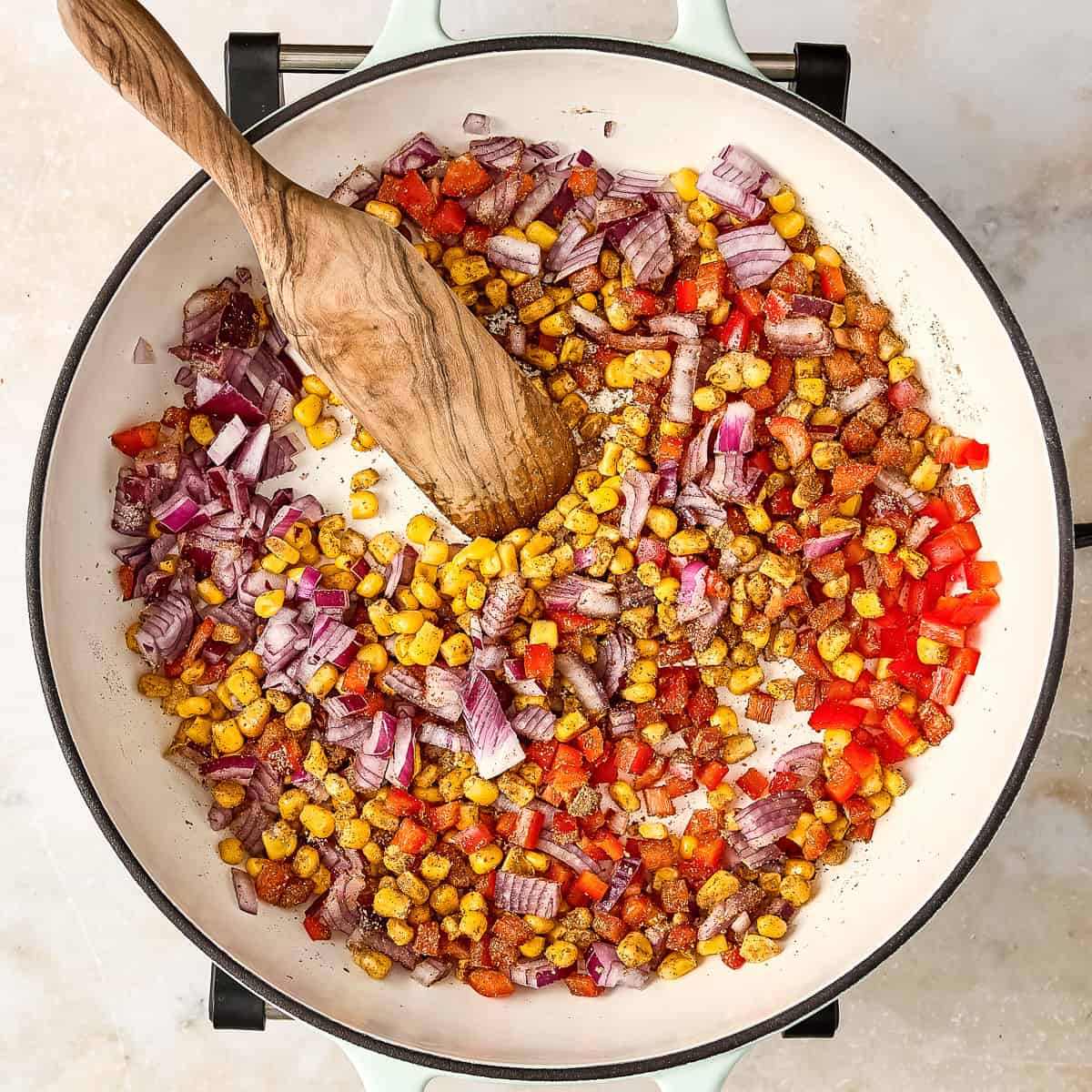 Sautéed peppers, onions, and corn cooking in a skillet with a wooden spoon.