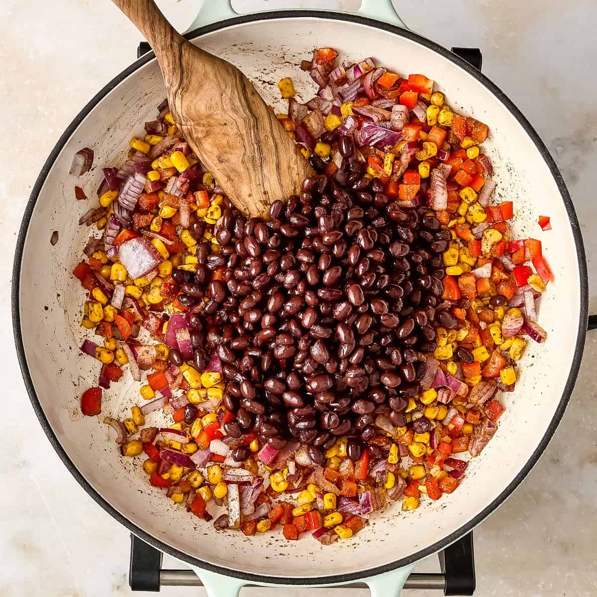 Black beans added to a skillet with cooked vegetables with a wooden spoon.