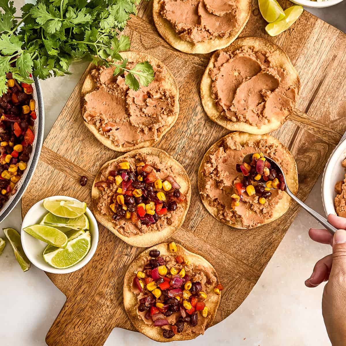 Woman adding cooked beans and veggies on top of refried beans on tostadas.