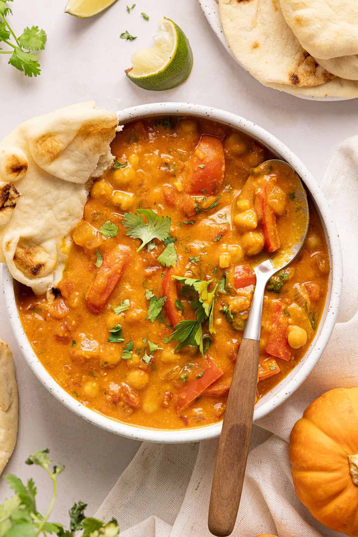 Bowl of pumpkin curry with a spoon and naan surrounded by ingredients.