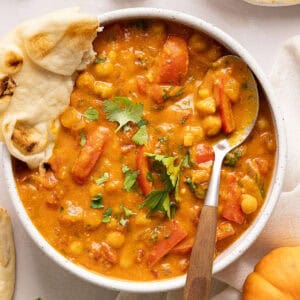 Bowl of pumpkin curry with a spoon and naan surrounded by ingredients.