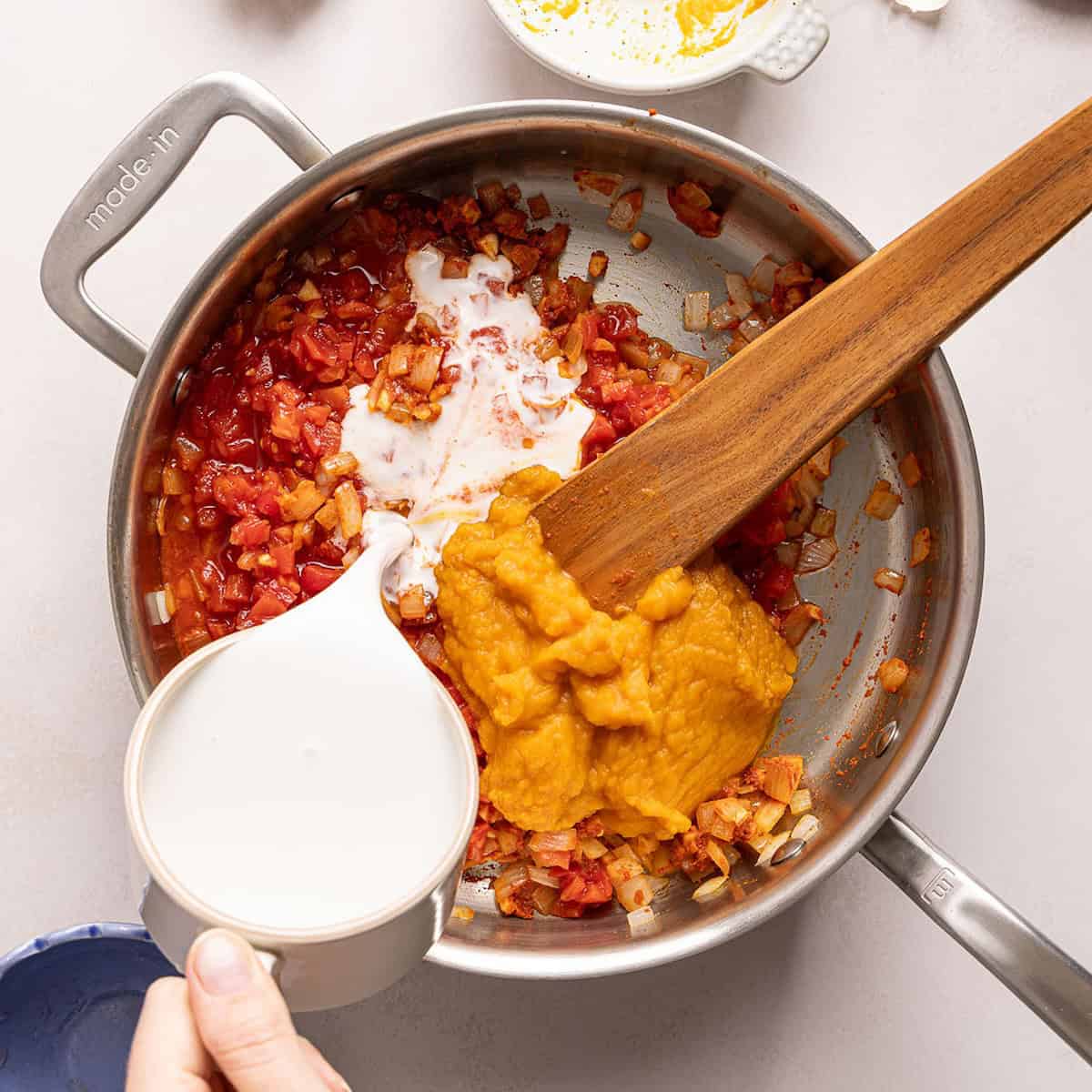 Woman pouring coconut milk into a skillet with onions, garlic, tomatoes, and pumpkin.