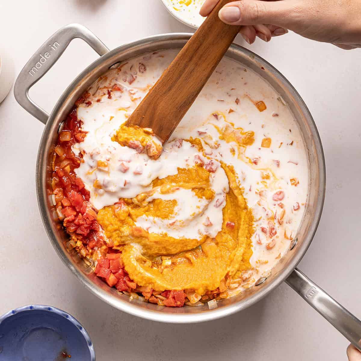 Woman stirring pumpkin, coconut milk, tomatoes, and onions in a large skillet.