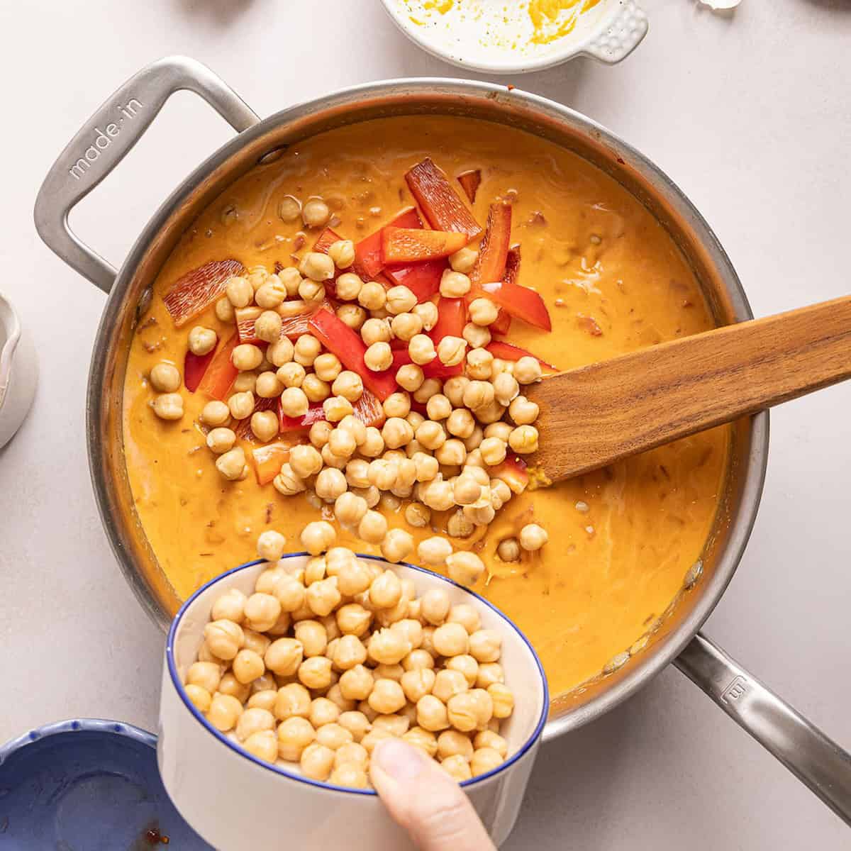 Woman adding chickpeas and red peppers to pumpkin curry in a skillet.