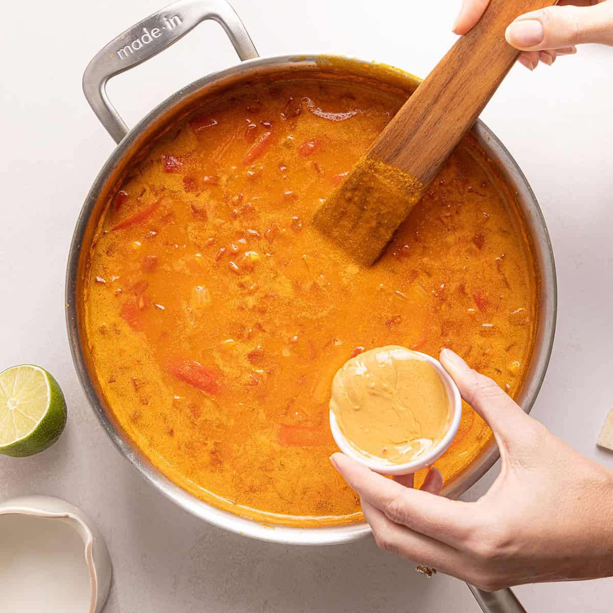 Woman pouring creamy peanut butter into pumpkin curry in a large skillet.