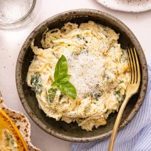 Spaghetti squash strands with healthy alfredo sauce in a bowl with a fork, cheese, and basil.