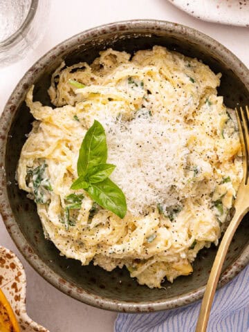Spaghetti squash strands with healthy alfredo sauce in a bowl with a fork, cheese, and basil.