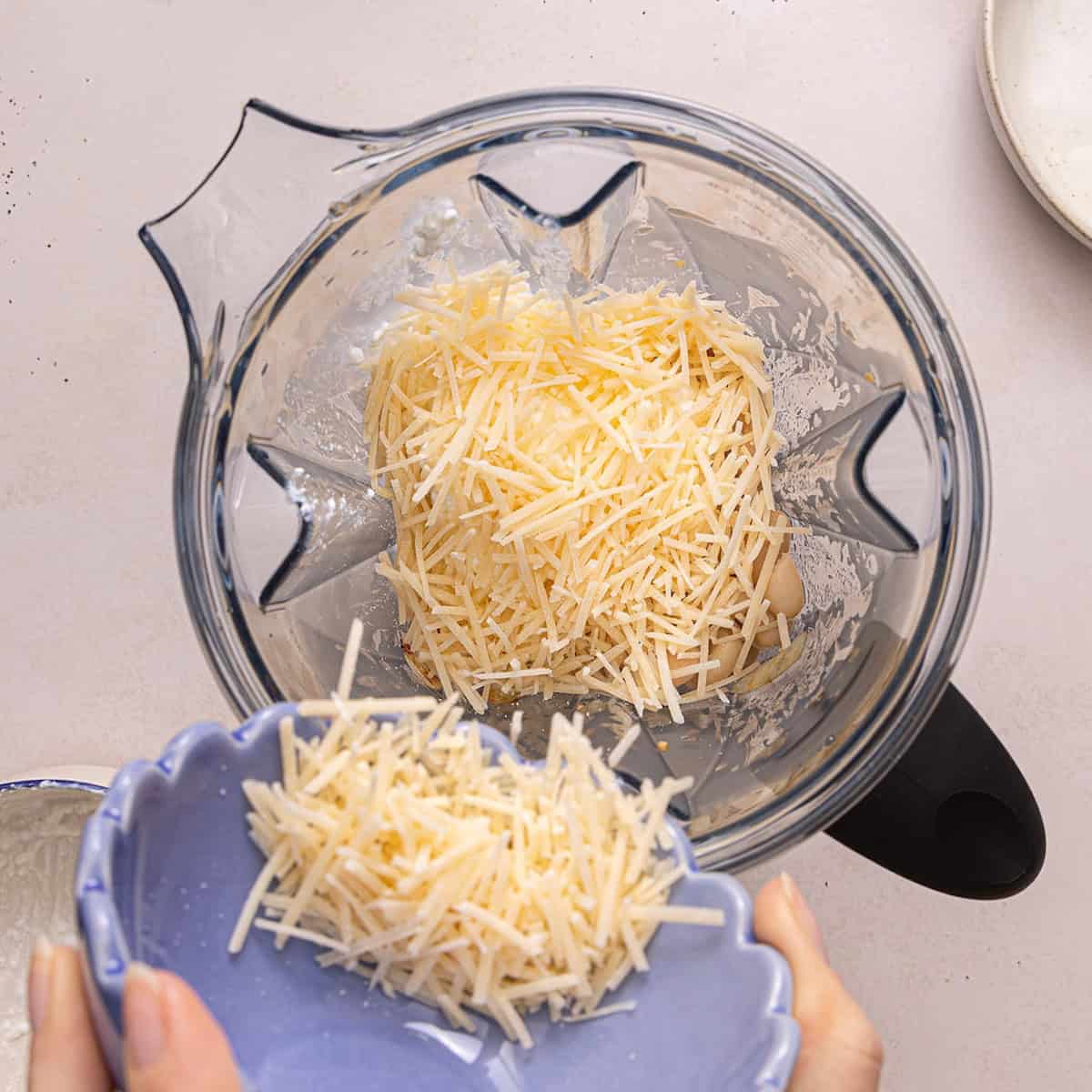 Woman adding parmesan cheese to a blender with other ingredients for alfredo.